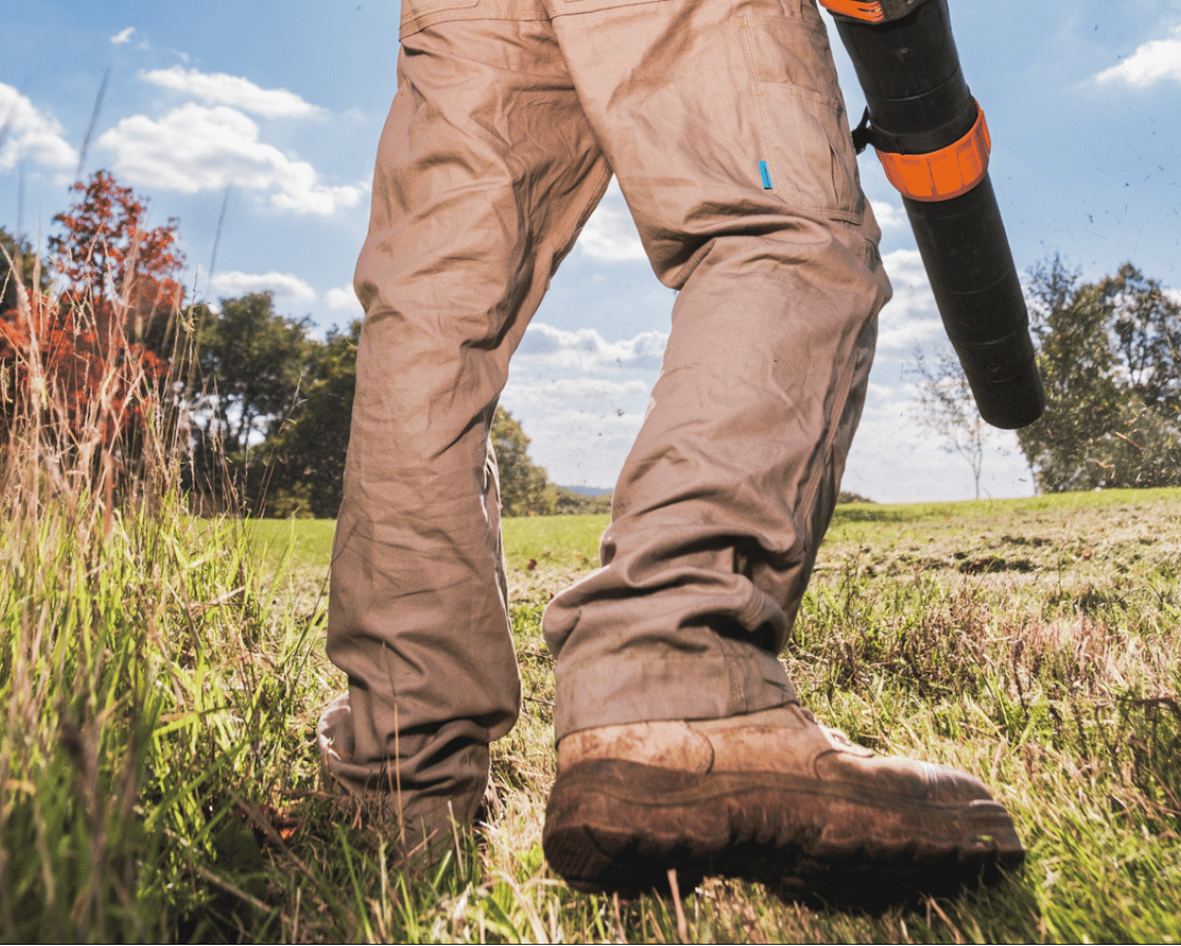 Person wearing brown work pants and boots holding a leaf blower in a grassy outdoor setting.